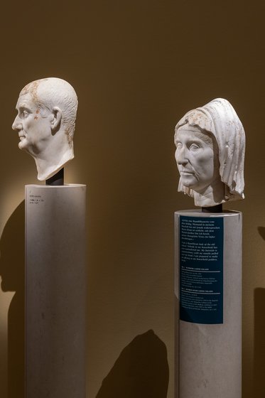 Zwei antike Porträtbüsten stehen auf Säulen. Links ist ein männliches Porträt mit kurzer Frisur und ernstem Ausdruck, rechts eine weibliche Büste mit Kopfdeckel und sanftem Gesichtsausdruck. Beide sind aus hellem Stein und vor einer neutralen Wand präsentiert.