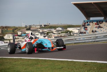Ein Rennwagen mit auffälligen blau-orangefarbenen Designelementen nimmt eine Kurve auf einer Rennstrecke. Im Hintergrund sind Zuschauer auf den Tribünen zu sehen. Die Szenerie vermittelt ein Gefühl von Geschwindigkeit und Spannung.