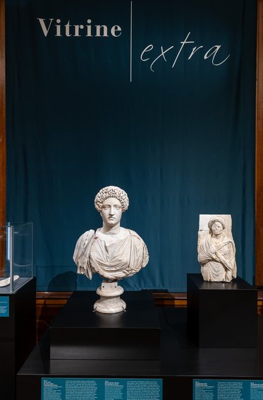 Eine Ausstellung mit zwei Skulpturen aus Marmor. Links steht eine Büste eines Mannes mit einer runden Frisur und einem Gewand. Rechts befindet sich eine kleinere Statue einer Frau, die ebenfalls ein Gewand trägt. Der Hintergrund ist blau und zeigt den Schriftzug "Vitrine | extra".