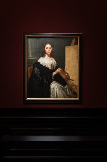 Ölbild einer Frau in historischer Kleidung, die an einem Tisch sitzt. Sie trägt ein schwarzes Gewand und hat lange, lockige Haare. Ihr Blick ist direkt auf den Betrachter gerichtet, während sie einen Pinsel in der Hand hält. Der Hintergrund ist dunkel und schlicht.