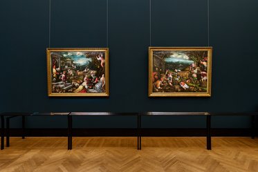 Zwei Gemälde in goldenen Rahmen hängen an einer dunkelgrünen Wand. Vor den Gemälden befindet sich eine lange, schlichte schwarze Vitrine. Der Boden ist aus hellem Holz, und der Raum wirkt wie eine Galerie oder ein Museum.