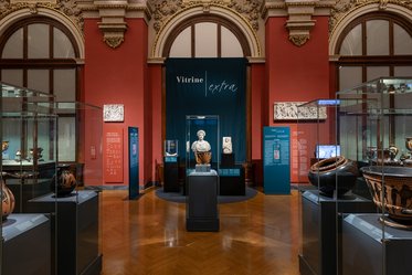 Ein Museumsraum mit rot-orangefarbenen Wänden. In der Mitte befindet sich eine Vitrine mit einer Statue. Rund um die Vitrine stehen mehrere Glaskästen mit antiken Vasen und anderen Artefakten. An den Wänden hängen Informationstafeln zu den Ausstellungsstücken.