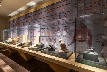 Der Bildausschnitt zeigt eine Museumsausstellung mit mehreren Vitrinen, die antike ägyptische Artefakte präsentieren. Im Hintergrund sind Wandmalereien mit Darstellungen von Menschen und Tieren zu sehen. Die Vitrinen enthalten Statuen und Skulpturen, die auf einer Holzplatte stehen.
