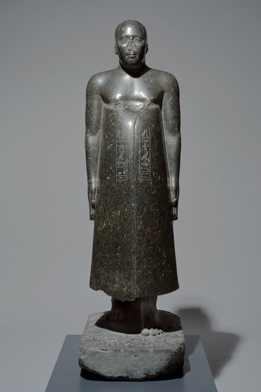 Eine aus schwarzem Stein gefertigte Statue eines menschlichen Körpers. Die Figur steht aufrecht, trägt ein langes Gewand und hat eine glatte Oberfläche. In der Mitte des Gewands sind hieroglyphische Inschriften eingraviert. Die Statue hat eine schlichte, aber imposante Erscheinung.