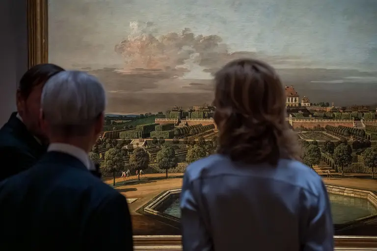 Two people stand in front of a large painting depicting a vast garden landscape with lush greenery, formal pathways, and a distant palace under a cloudy sky. The individuals are seen from behind, engaged in observation of the artwork.