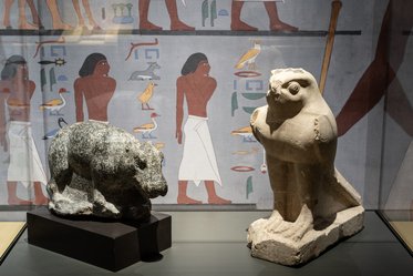 Zwei antike ägyptische Statuen sind zu sehen: Links ein steinerner Löwe, rechts eine Statue eines geflügelten Wesens. Im Hintergrund befindet sich eine Wandmalerei mit Szenen aus dem alten Ägypten und roten sowie grünen Symbolen.