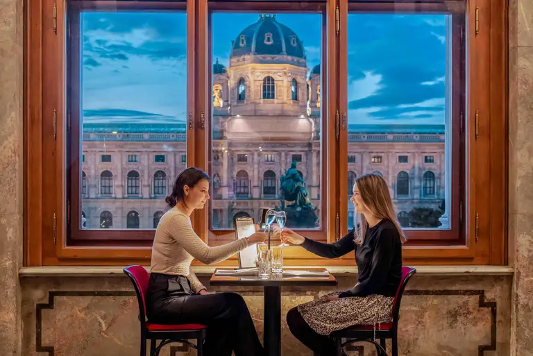 Zwei Frauen sitzen an einem Tisch in einem Restaurant mit Blick auf ein historisches Gebäude. Sie stoßen mit Gläsern an und scheinen ein angenehmes Gespräch zu führen. Der Hintergrund zeigt eine Abendskyline mit einem bewölkten Himmel.