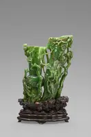Eine kunstvolle Skulptur aus grünem Jade, die eine natürliche Szenerie mit Pflanzen und ergänzt durch reliefartige Figurenelemente darstellt. Die Skulptur steht auf einem dekorativen Holzsockel.
