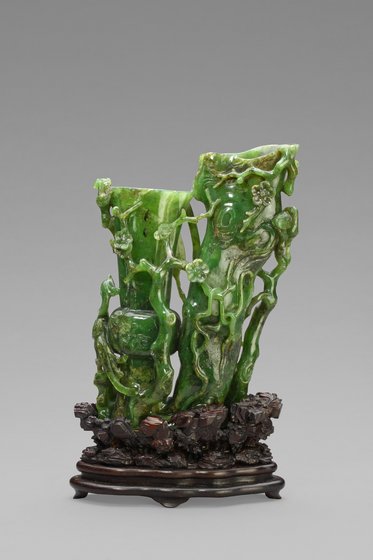 Eine kunstvolle Skulptur aus grünem Jade, die eine natürliche Szenerie mit Pflanzen und ergänzt durch reliefartige Figurenelemente darstellt. Die Skulptur steht auf einem dekorativen Holzsockel.