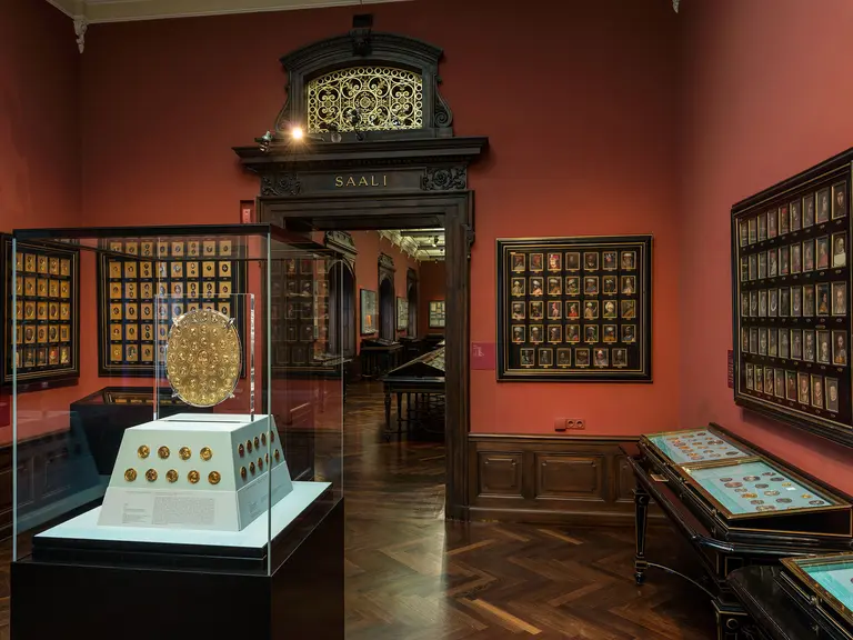 Ein Innenraum eines Museums mit roten Wänden. Im Vordergrund steht ein vitrinenartige Ausstellungstisch mit einer runden Goldscheibe und mehreren weiteren Objekten. An den Wänden hängen zahlreiche Bilderrahmen mit Porträts. Der Eingang zu einem anderen Raum ist sichtbar.