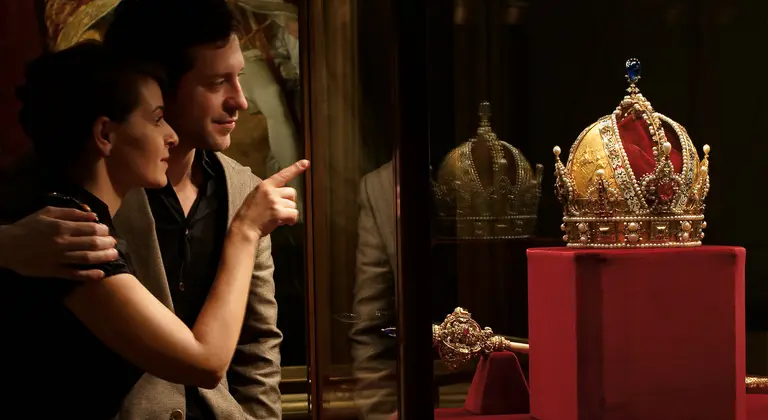 A couple admires an ornate crown displayed in a glass case. The crown, embellished with jewels and intricate designs, sits on a red pedestal. The woman points at the crown while the man looks on, both showcasing a moment of curiosity and engagement.