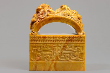 Das Bild zeigt einen kunstvoll geschnitzten jadefarbenen Stein, der mit einem geschwungenen oberen Teil und detaillierten Reliefs verziert ist. Das Material hat eine gelbliche Farbe, und die Motive sind von traditioneller asiatischer Kunst inspiriert.