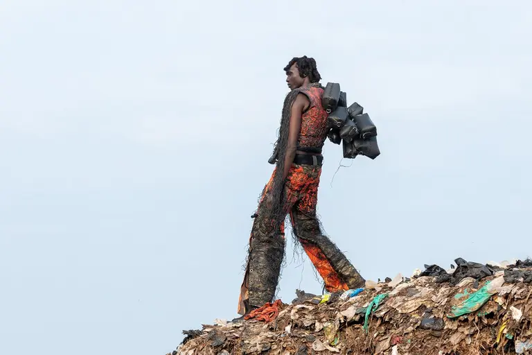A person walks on a large pile of waste, wearing an outfit made of colorful, discarded materials. The background features a cloudy sky, emphasizing the contrast between the individual and the surrounding garbage.
