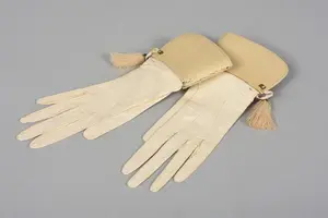 Das Bild zeigt ein Paar elegante, cremefarbene Lederhandschuhe mit einem dekorativen, gefalteten Abschluss. Die Handschuhe sind länglich und haben eine feine, geschmeidige Textur. An einem der Abschlüsse hängt eine kleine Quaste als Verzierungen.