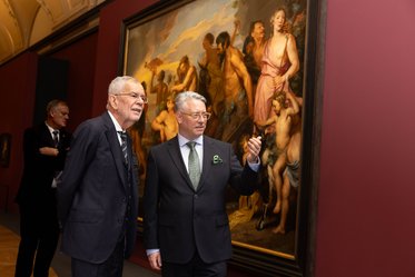 Zwei Männer in Anzügen stehen vor einem großen Gemälde, das eine historische oder mythologische Szene zeigt. Der Mann auf der linken Seite lächelt und spricht, während der andere aufmerksam zuhört. Im Hintergrund sind weitere Personen und Teile des Museums zu sehen.