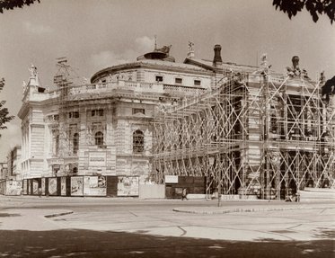 Das Bild zeigt ein historisches Gebäude, das derzeit renoviert wird. Gerüste umgeben die Fassade, während Arbeiten im Gange sind. Im Vordergrund befinden sich Plakate, die auf das Theater hinweisen. Die Aufnahme ist in sepia gehalten, was eine nostalgische Atmosphäre schafft.