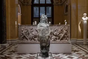 Eine Skulptur eines Mannes mit einer beschädigten Oberfläche steht vor einem reliefierten Hintergrund, der antike Szenen zeigt. Die Statue ist in einem Museum aufgestellt, umgeben von eleganter Architektur und weiteren Kunstwerken.