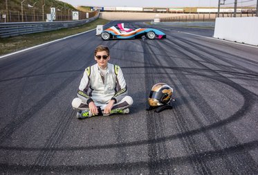Ein junger Rennfahrer in Rennanzug sitzt im Schneidersitz auf der Rennstrecke, neben seinem Helm. Im Hintergrund ist ein Rennwagen zu sehen, der sich mit Reifenabdrücken in einer Kurve bewegt. Die Szenerie vermittelt ein Gefühl von Geschwindigkeit und Motorsport.