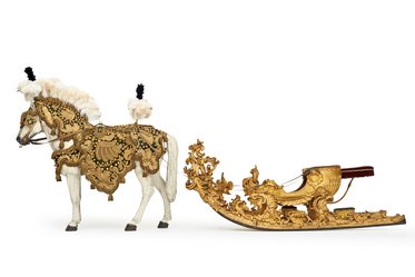 Ein detailliert verziertes, weißes Holzpferd mit Federn und prächtigen Ornamenten zieht eine goldene, kunstvoll gestaltete Schlitten. Beide Objekte zeigen aufwändige Schnitzereien und sind historisch inspiriert.