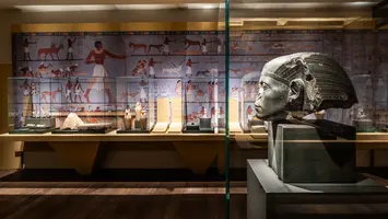 In einem Museum sind verschiedene ägyptische Artefakte ausgestellt. Im Vordergrund steht eine große Kopfskulptur. Im Hintergrund sind Wände mit Wandmalereien und Vitrinen, die weitere historische Objekte zeigen. Das Licht beleuchtet die Exponate und schafft eine interessante Atmosphäre.