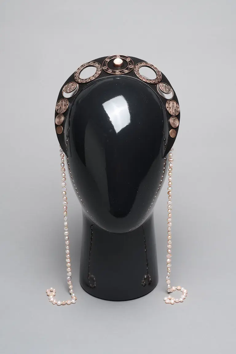 A decorative headpiece displayed on a black mannequin head. The headpiece features intricate designs with circular elements and is adorned with strings of pearls hanging down the sides. The background is a neutral gray, highlighting the details of the accessory.