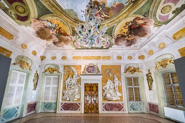 Bild eines prächtigen Raumes mit einem kunstvoll bemalten Deckengewölbe, einer eleganten Glasleuchte und dekorativen Wänden. An den Wänden sind Skulpturen und farbige Fresken zu sehen, die historische Szenen darstellen. Die Tür ist zentral im Raum angeordnet.