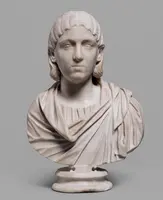 Eine antike Marmorbüste eines jungen Mannes mit langen, lockigen Haaren. Er trägt ein gefaltetes Gewand und hat einen neutralen Gesichtsausdruck. Die Details der Kleidung und der Gesichtszüge sind gut ausgearbeitet. Die Statue steht auf einem runden Sockel.