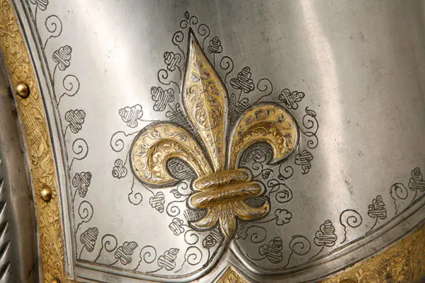Das Bild zeigt ein metallisches Rüstungsteil mit kunstvollem Fleur-de-Lis-Motiv, umgeben von geschwungenen Mustern. Das Design ist in Silber und Gold gehalten und vermittelt einen Eindruck von historischer Handwerkskunst und Eleganz.