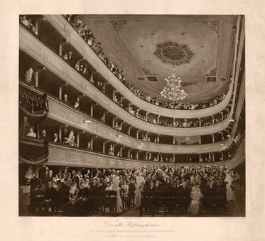 Das Bild zeigt den Innenraum eines alten Theaters mit mehreren Rängen und einer großen Menschenmenge. Die Zuschauer blicken nach vorne, wo eine Aufführung stattfindet. Eine eindrucksvolle Deckenleuchte und aufwendige Dekorationen heben die Atmosphäre des Theaters hervor.