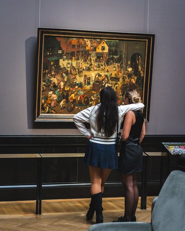 Zwei Frauen stehen vor einem Gemälde in einem Museum und betrachten es. Die eine Frau hat lange, dunkle Haare und trägt einen weißen Pullover und einen blauen Rock, die andere hat kurze, blonde Haare und trägt ein schwarzes Outfit.