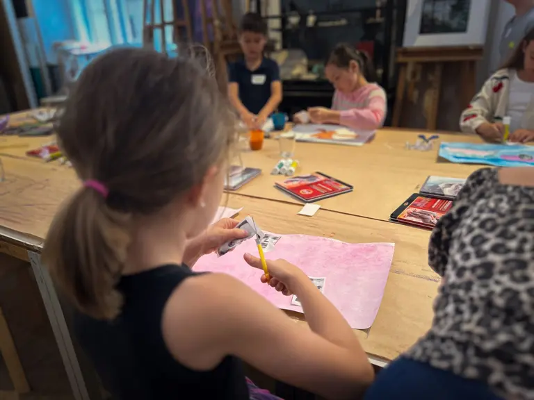 Eine Gruppe von Kindern, die an einem Tisch sitzt und kreativ arbeitet. Ein Mädchen in schwarz schneidet mit einer Schere ein Stück Papier auf einer rosa Unterlage. Im Hintergrund sind weitere Kinder mit verschiedenen Bastelmaterialien beschäftigt.