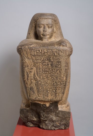Eine antike ägyptische Statue aus Stein, die eine menschliche Figur mit einer schmalen, ausdrucksvolle Gesicht zeigt. Die Figur hält ein Schriftstück mit Hieroglyphen, die auf ihrer Vorderseite eingraviert sind. Die Statue steht auf einem roten Sockel.