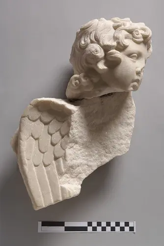 Eine detailreiche, marmorene Skulptur eines ängstlichen Kopfes mit Locken und einem Flügel. Der Kopf zeigt einen nachdenklichen Ausdruck, während er leicht zur Seite geneigt ist. Die Mühe der Kunstfertigkeit ist in den feinen Details der Haare und Flügel sichtbar.