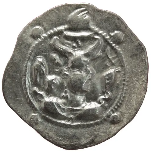 Eine silberne Münze zeigt eine stilisierte Darstellung von Figuren in einem kreisförmigen Design. Eine Figur trägt einen Hut und es sind zusätzliche Details im Hintergrund sichtbar. Die Münze hat einen unregelmäßigen Rand und eine matte Oberfläche.