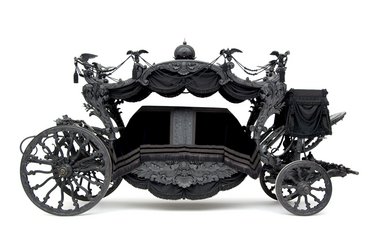 Eine schwarze, kunstvoll verzierte Leichentrage mit offenen Seiten zeigt einen eleganten Innenraum. Die Räder sind kunstvoll gestaltet, und das gesamte Fahrzeug strahlt eine düstere, aber opulente Ästhetik aus.