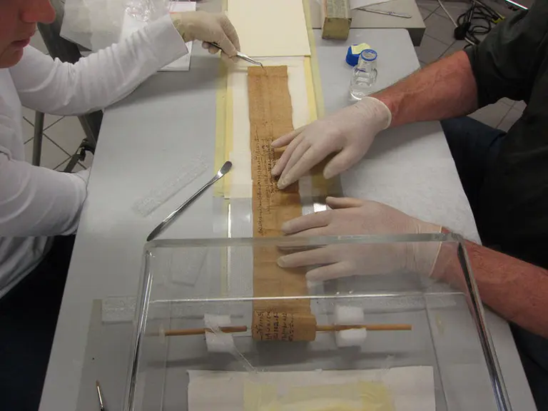 Zwei Personen arbeiten an der Konservierung eines alten Manuskripts. Der Textstreifen liegt auf einem Tisch, während eine Person mit einer Pinzette und die andere mit der Hand das Manuskript vorsichtig behandelt. Schutzhandschuhe werden getragen, um das Dokument zu schützen.