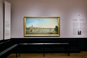 Ein Gemälde in einem Museum zeigt einen Hafen mit Booten und einer Stadtansicht im Hintergrund. Der Rahmen des Gemäldes ist goldfarben. An der Wand neben dem Bild sind erklärende Texte und Informationen über die Ausstellung angebracht.