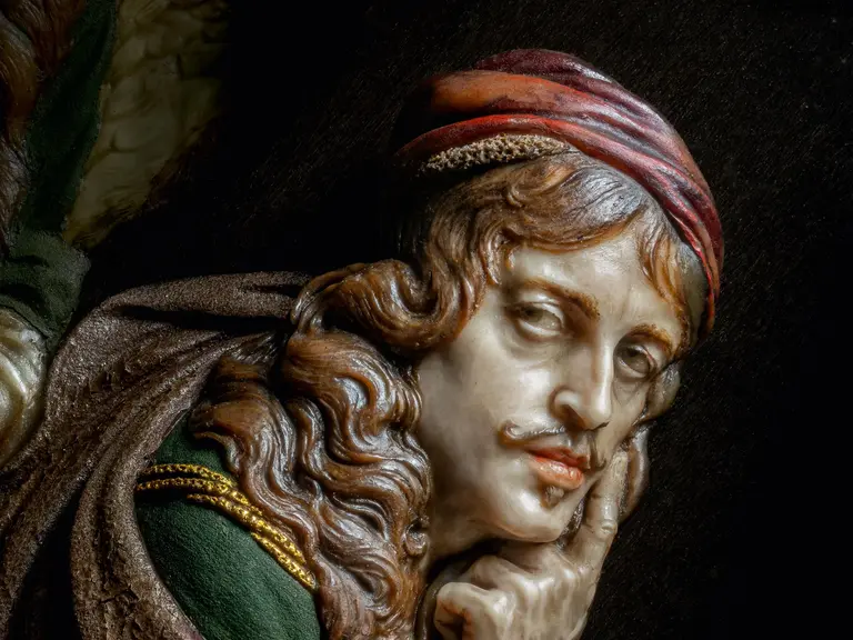 A wax sculpture by Daniel Neuberger of a detailed head of a man with curly hair and a red headscarf. He has a thoughtful expression on his face and is holding a hand to his chin. His robe is green with gold accents and the background is dark. 