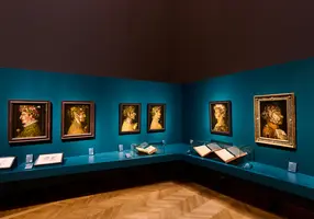 Im Bild ist eine Galerie mit mehreren Portraitgemälden zu sehen, die an einer türkisfarbenen Wand hängen. Die Gemälde zeigen Personen mit verschiedenen Kopfbedeckungen, darunter Blumen und Laub. Auf einem Tisch davor liegen einige Bücher, deren Seiten teilweise geöffnet sind.