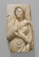 Reliefdarstellung einer Figur in antiker Kleidung, mit einem drapierten Umhang und lockigem Haar. Die Gesichtszüge sind teilweise abgetragen. Die Figur steht frontal und hält eine Hand an ihrem Oberkörper. Der Hintergrund ist schlicht.