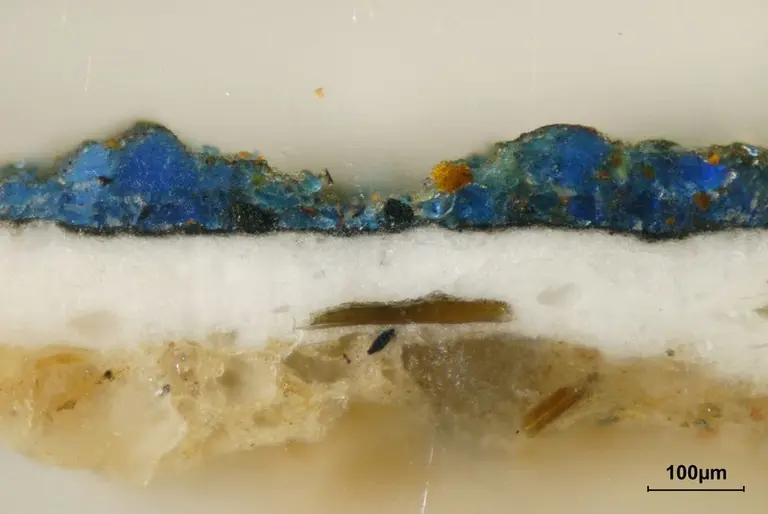 Mikroskopisches Bild eines Gesteins mit mehreren Schichten. Die obere Schicht enthält leuchtend blaue Mineralien, während die darunterliegende Schicht weiß und sandig ist. Eine darunterliegende Schicht ist ebenfalls sichtbar. Eine Maßstabsmarke von 100 Mikrometern ist im Bild enthalten.
