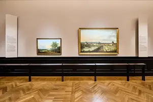 Zwei Gemälde in einer Galerie. Links ein kleineres Landschaftsbild mit einem Baum, rechts ein größeres Stadtansichtsgemälde. Im Hintergrund stehen Informationsschilder, und der Boden ist aus Holz mit einem auffälligen Muster.