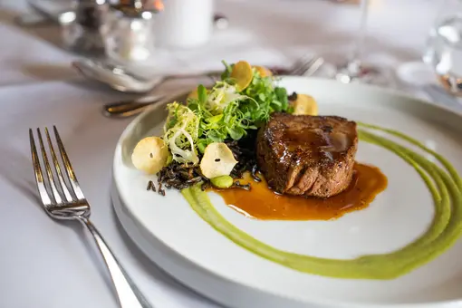 Ein Teller mit saftigem Fleisch, umgeben von einer Sauce, serviert auf einem Bett aus schwarzem Reis. Daneben liegt frischer Blattsalat und knusprige Chips. Die Anrichtung ist ansprechend mit einem grünen Püree.