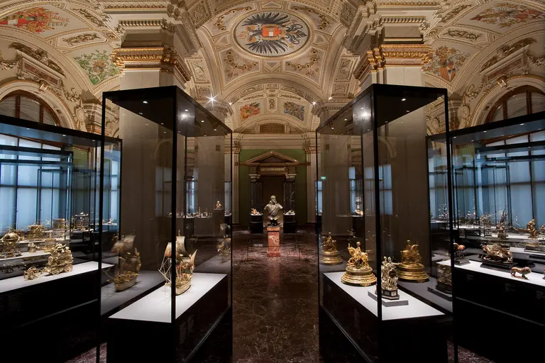 Im Bild ist ein prunkvoller Museumssaal zu sehen, in dem Vitrinen mit kunstvollen Objekten aufgestellt sind. Die Decke ist reich verziert, und die Beleuchtung hebt die ausgestellten Stücke hervor. Der Raum vermittelt eine Atmosphäre von kulturellem Erbe und Geschichte.