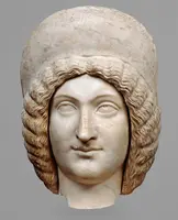 Eine antike Marmorbüste mit einem weiblichen Gesicht zeigt eine sanfte Ausdrücklichkeit. Die Frisur ist wellig und reich verziert, während die Kopfpartie von einem einfachen Kopfschmuck umrahmt wird. Die Augen und der Mund sind leicht geöffnet, was einen erkennbaren, nachdenklichen Ausdruck verleiht.