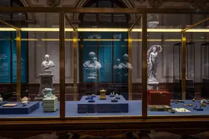 Ein Ausstellungsraum mit Vitrinen, die verschiedene historische Artefakte zeigen. Darunter sind Statuen und kleine Objekte, umgeben von informativen Hintergründen. Im Hintergrund sind weitere Skulpturen sichtbar, die die Tiefe des Raumes betonen.
