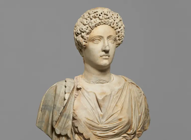 Eine antike Marmorstatue einer Frau mit lockigem Haar und einem gefalteten Gewand. Die Statue zeigt den Oberkörper und ist detailreich gearbeitet, insbesondere im Gesicht und der Kleidung. Der Hintergrund ist neutral grau.