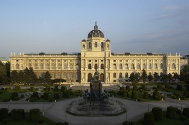 Das Bild zeigt die prächtige Fassade des Kunsthistorischen Museums in Wien, umgeben von gepflegten Gärten und Bäumen. Im Vordergrund steht ein Denkmal, das die Aufmerksamkeit auf die imposante Architektur und den geschmackvollen Landschaftsgestaltung lenkt.