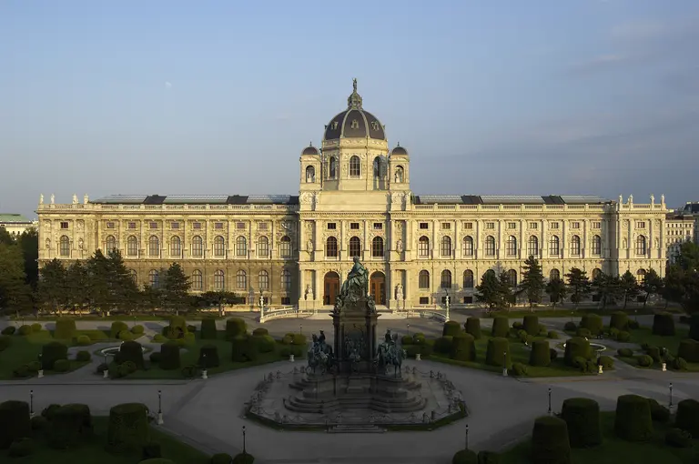 Das Bild zeigt die prächtige Fassade des Kunsthistorischen Museums in Wien, umgeben von gepflegten Gärten und Bäumen. Im Vordergrund steht ein Denkmal, das die Aufmerksamkeit auf die imposante Architektur und den geschmackvollen Landschaftsgestaltung lenkt.