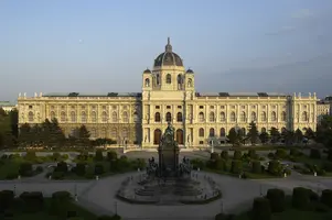 Das Bild zeigt die prächtige Fassade des Kunsthistorischen Museums in Wien, umgeben von gepflegten Gärten und Bäumen. Im Vordergrund steht ein Denkmal, das die Aufmerksamkeit auf die imposante Architektur und den geschmackvollen Landschaftsgestaltung lenkt.